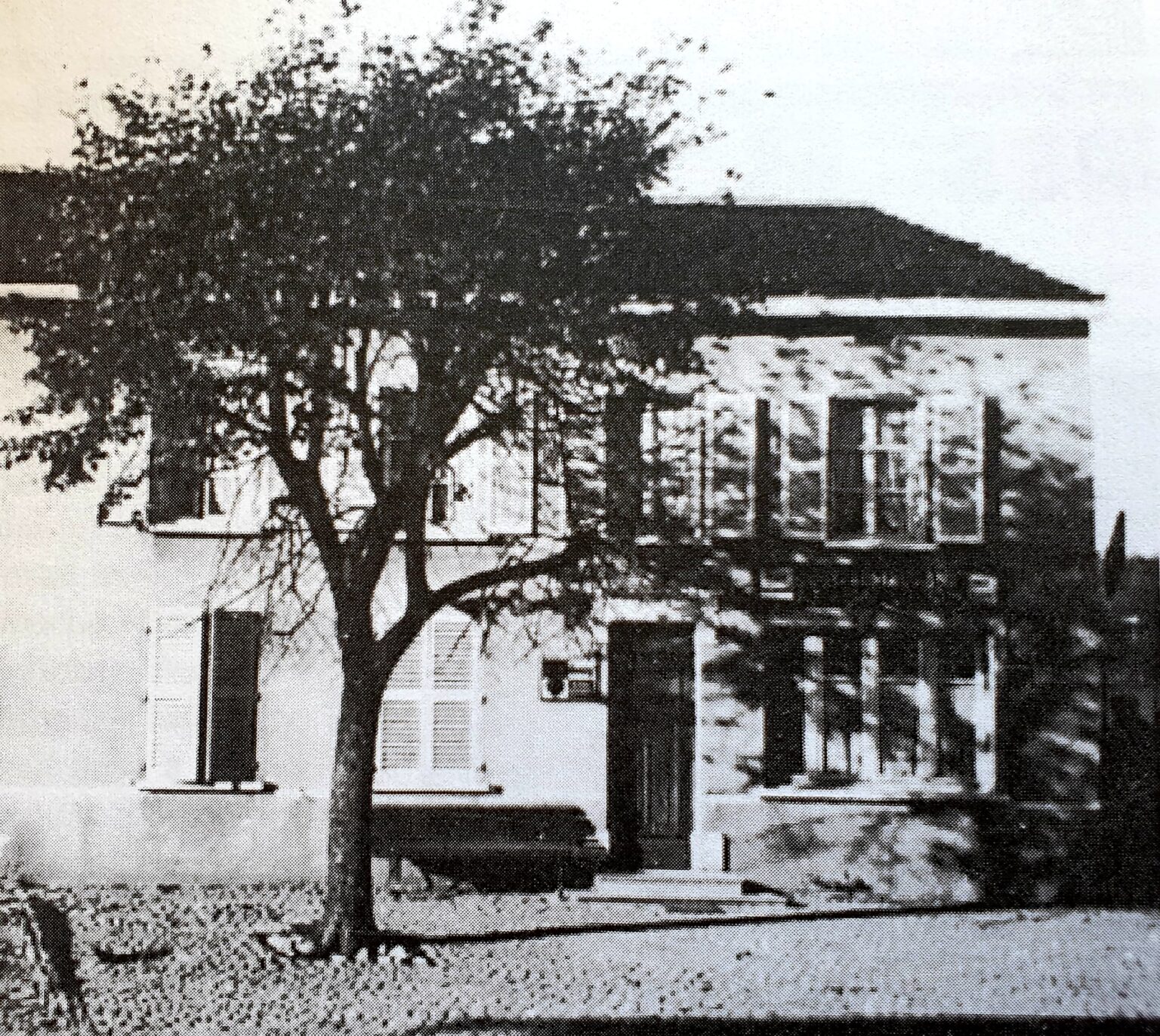 Une grande maison derrière un arbre au feuillage épais, avec une terrasse couverte à l'étage et des volets de bois ; une voiture est garée devant l'entrée.