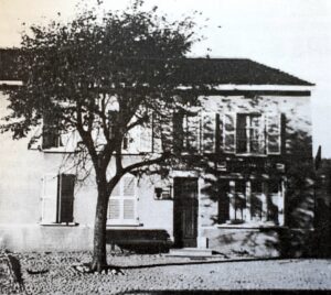 Une grande maison derrière un arbre au feuillage épais, avec une terrasse couverte à l'étage et des volets de bois ; une voiture est garée devant l'entrée.