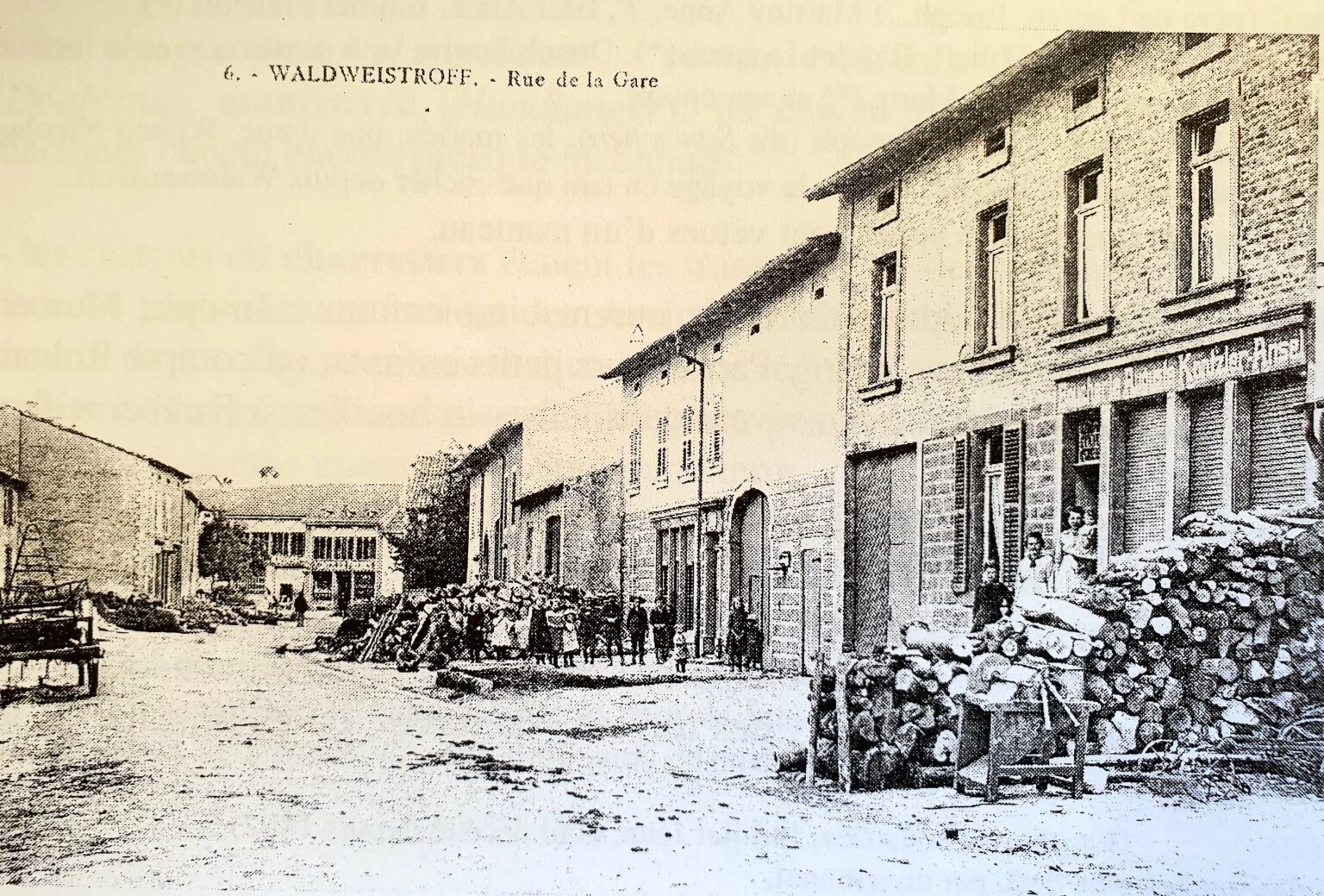 Carte postale de la « Rue de la Gare » : une large rue bordée de maisons et d'énormes tas de bois de chauffage ; un groupe d'enfants et d'adultes pose au milieu de la chaussée.