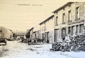 Carte postale de la « Rue de la Gare » : une large rue bordée de maisons et d'énormes tas de bois de chauffage ; un groupe d'enfants et d'adultes pose au milieu de la chaussée.