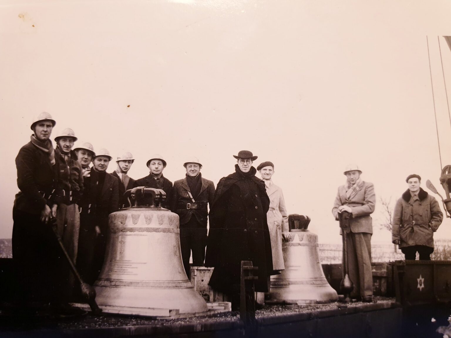 Une douzaine d'hommes, dont plusieurs pompiers casqués et un prêtre en manteau noir et chapeau, posent derrière deux grosses cloches posées sur un wagon.