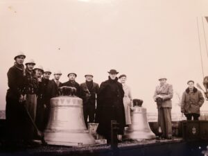 Une douzaine d'hommes, dont plusieurs pompiers casqués et un prêtre en manteau noir et chapeau, posent derrière deux grosses cloches posées sur un wagon.