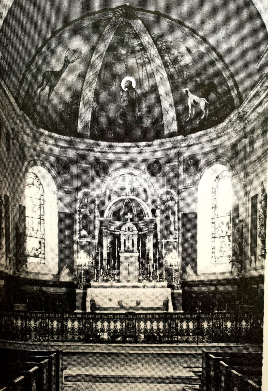 Le chœur de l'église : la coupole peinte montre un saint agenouillé en forêt entre un cerf et des chiens ; au-dessous, le maître-autel garni de chandeliers et la table de communion en fer forgé.
