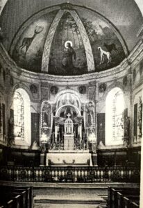 Le chœur de l'église : la coupole peinte montre un saint agenouillé en forêt entre un cerf et des chiens ; au-dessous, le maître-autel garni de chandeliers et la table de communion en fer forgé.
