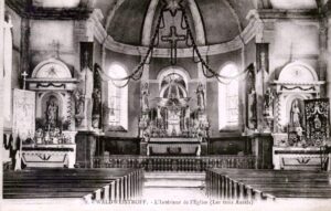 Carte postale ancienne de l'intérieur de l'église : les trois autels, les statues et les bannières, des guirlandes tendues sous la voûte, et les bancs de bois au premier plan.