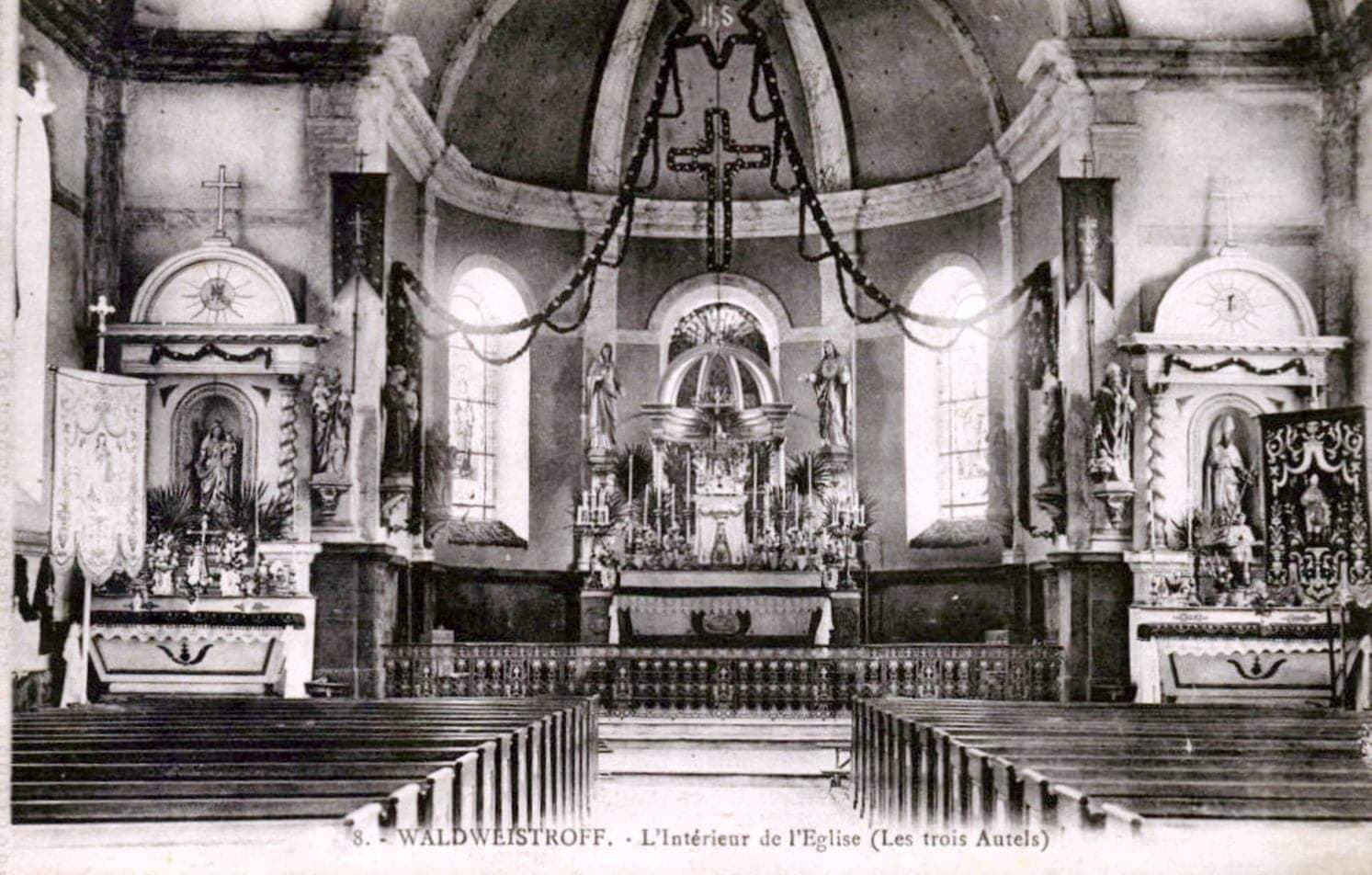 Carte postale ancienne de l'intérieur de l'église : les trois autels, les statues et les bannières, des guirlandes tendues sous la voûte, et les bancs de bois au premier plan.