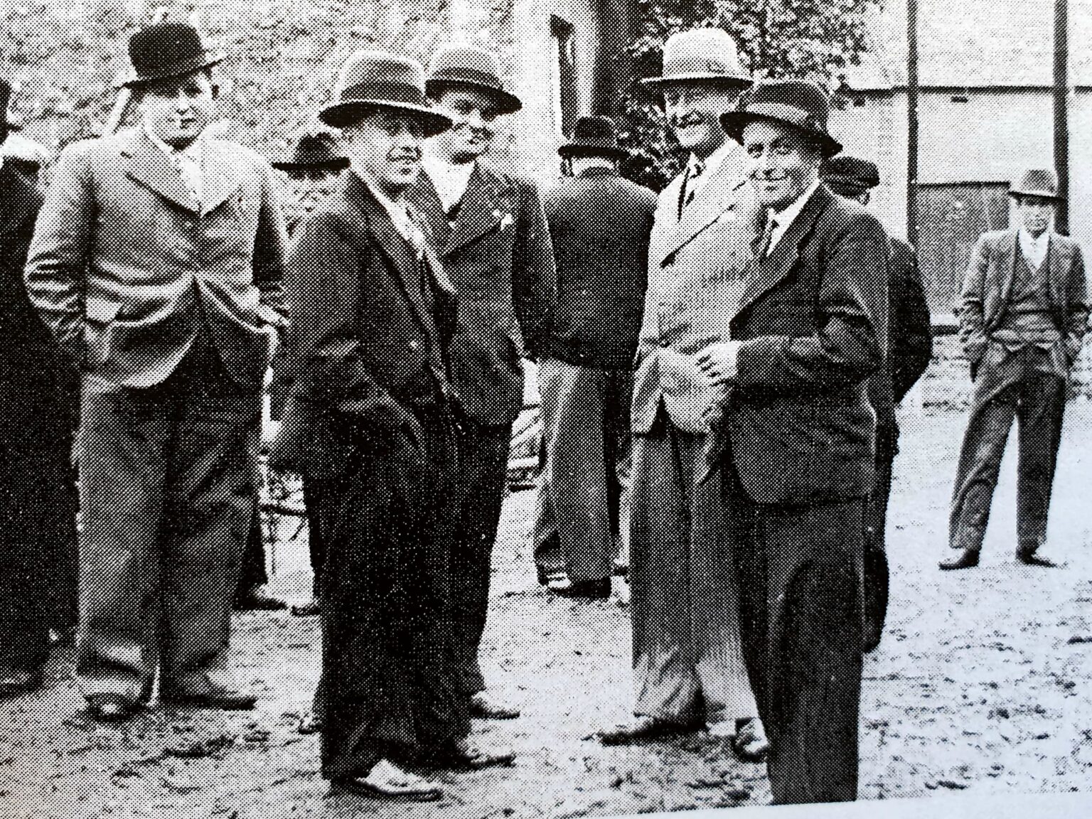 Une dizaine d'hommes en chapeau et manteau discutent debout sur une place, les mains dans les poches ; un autre s'éloigne en arrière-plan.
