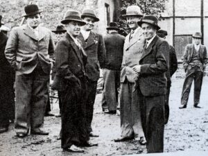 Une dizaine d'hommes en chapeau et manteau discutent debout sur une place, les mains dans les poches ; un autre s'éloigne en arrière-plan.