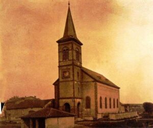 L'église vue de l'extérieur : une haute tour à flèche pointue portant une horloge, la nef percée de fenêtres en plein cintre, et les maisons basses du village à gauche.