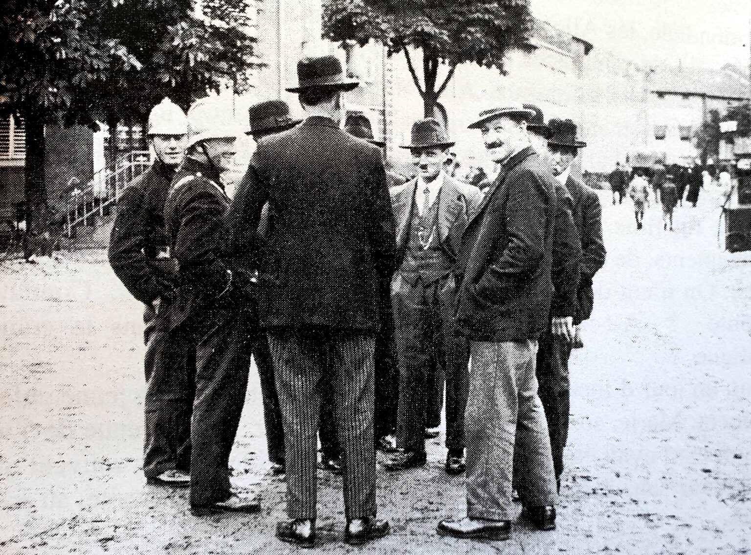 Des hommes en chapeau et manteau discutent sur la place du village, deux sapeurs-pompiers en casque blanc parmi eux ; des arbres et les maisons ferment le fond.