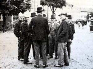Des hommes en chapeau et manteau discutent sur la place du village, deux sapeurs-pompiers en casque blanc parmi eux ; des arbres et les maisons ferment le fond.