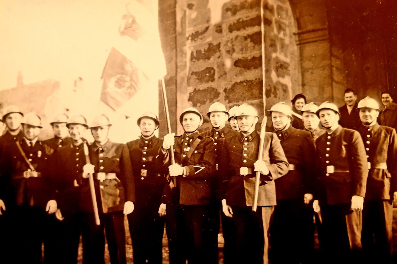 Une quinzaine de sapeurs-pompiers en casque et en uniforme, certains tenant une hache ou une pique, alignés devant le portail de l'église sous deux drapeaux.