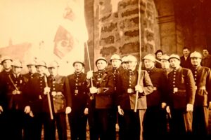 Une quinzaine de sapeurs-pompiers en casque et en uniforme, certains tenant une hache ou une pique, alignés devant le portail de l'église sous deux drapeaux.