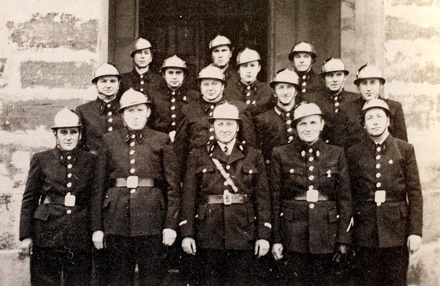 Portrait de groupe : une quinzaine de sapeurs-pompiers en casque et vareuse à boutons, sur trois rangs devant la porte d'un bâtiment.