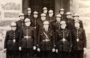 Portrait de groupe : une quinzaine de sapeurs-pompiers en casque et vareuse à boutons, sur trois rangs devant la porte d'un bâtiment.