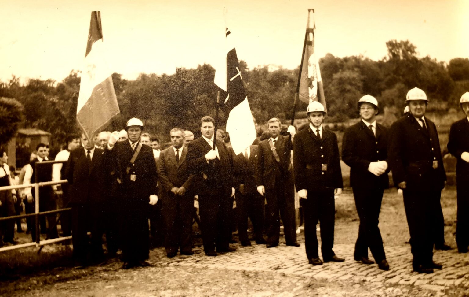 Rassemblement en plein air : trois drapeaux dont le tricolore, des hommes en costume et des sapeurs-pompiers en casque blanc alignés sur un sol pavé, le public derrière une barrière.