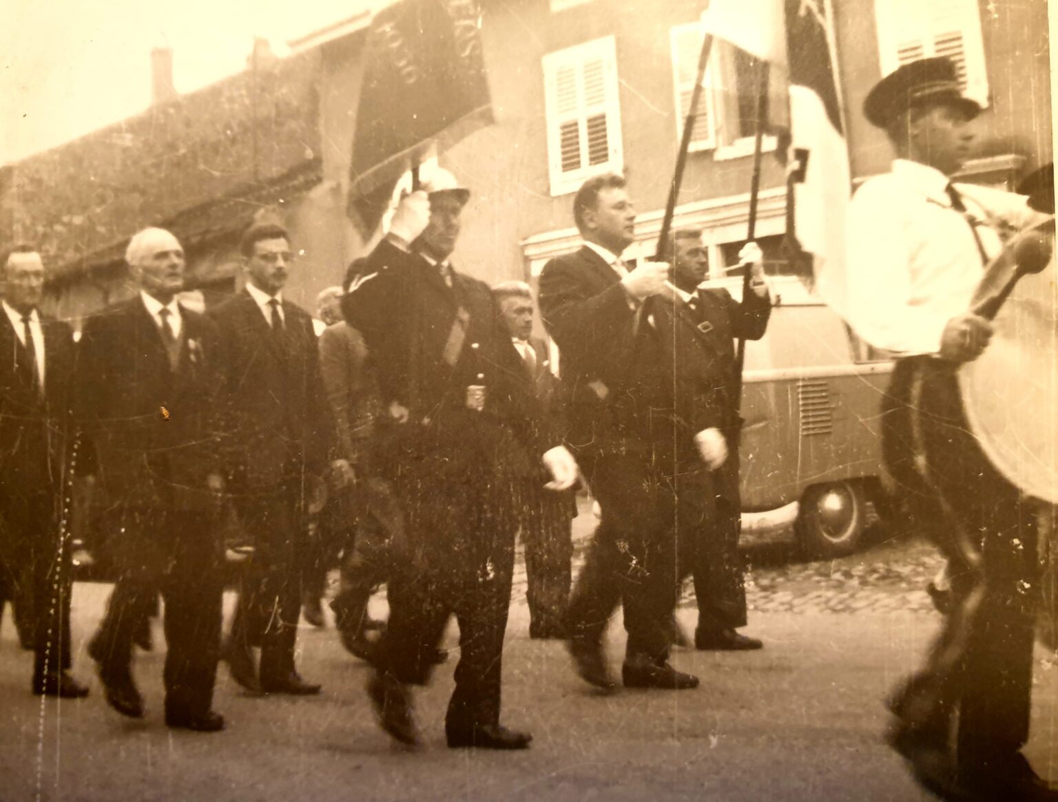 Cortège d'hommes en costume et de sapeurs-pompiers derrière une bannière ; à droite, un tambour en chemise blanche bat la marche, et une camionnette est garée le long des maisons.