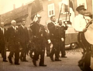 Cortège d'hommes en costume et de sapeurs-pompiers derrière une bannière ; à droite, un tambour en chemise blanche bat la marche, et une camionnette est garée le long des maisons.