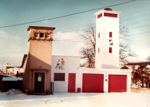 Sous la neige, le local des pompiers : une tour blanche surmontée d'une sirène, trois portes de garage rouges, une fresque murale représentant deux enfants, et l'ancienne tour du transformateur à gauche.