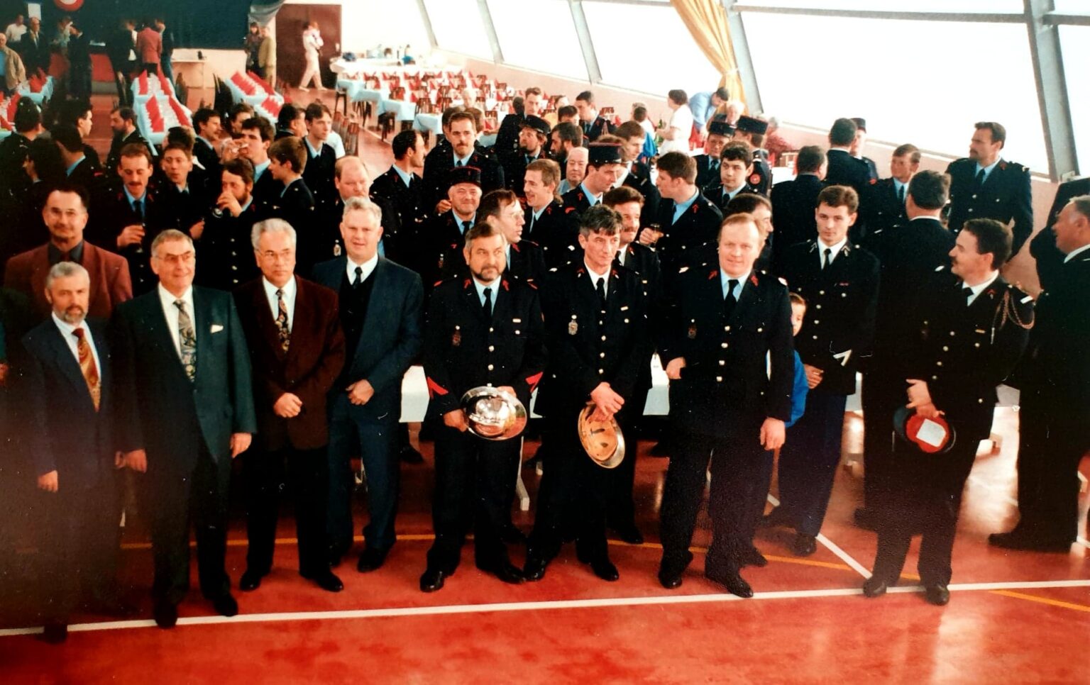 Cérémonie dans une salle au sol rouge : des sapeurs-pompiers en tenue de sortie, casques à la main, entourés d'élus en costume ; derrière eux, des tables dressées et une assistance nombreuse.