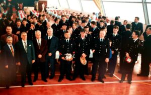Cérémonie dans une salle au sol rouge : des sapeurs-pompiers en tenue de sortie, casques à la main, entourés d'élus en costume ; derrière eux, des tables dressées et une assistance nombreuse.