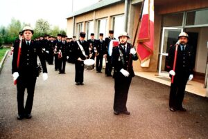Formation de sapeurs-pompiers devant un bâtiment moderne : un porte-drapeau au centre, un tambour et une clique de clairons, et deux rangs alignés derrière.