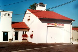 Le local des pompiers : un bâtiment blanc à toit de tuiles rouges surmonté d'une tour de séchage, l'inscription « Sapeurs Pompiers » en lettres rouges au-dessus de la porte du garage, et des bacs de fleurs devant l'entrée.