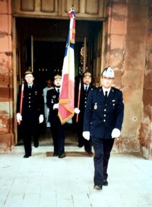 Des sapeurs-pompiers en tenue de sortie sortent du portail de l'église : l'un porte le drapeau tricolore, trois autres des casques de cuivre et des bâtons rouges, celui qui ouvre la marche un casque argenté.