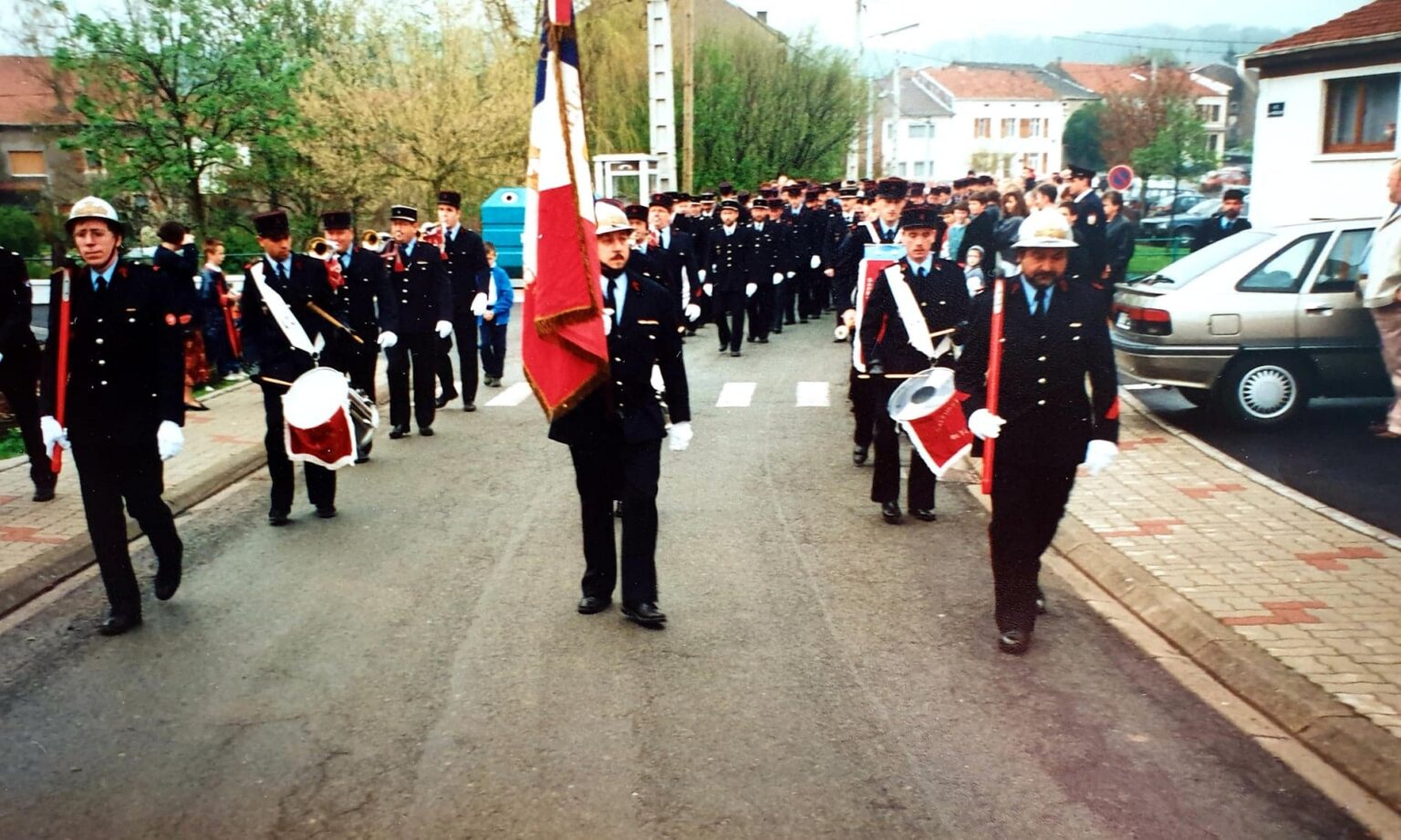 Défilé dans la rue : un porte-drapeau en tête, des tambours et des clairons de chaque côté, et une longue colonne de sapeurs-pompiers en uniforme suivie par les habitants.