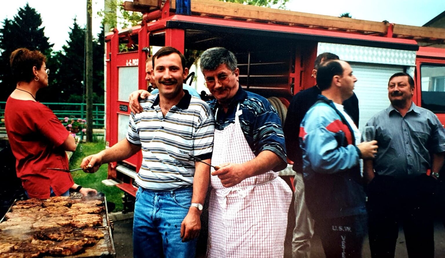 Barbecue devant un engin de secours rouge : deux hommes, dont l'un en tablier blanc, tiennent des brochettes au-dessus d'une grille couverte de viande, entourés d'autres convives.