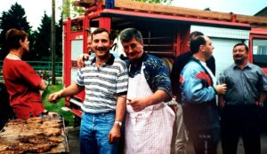 Barbecue devant un engin de secours rouge : deux hommes, dont l'un en tablier blanc, tiennent des brochettes au-dessus d'une grille couverte de viande, entourés d'autres convives.