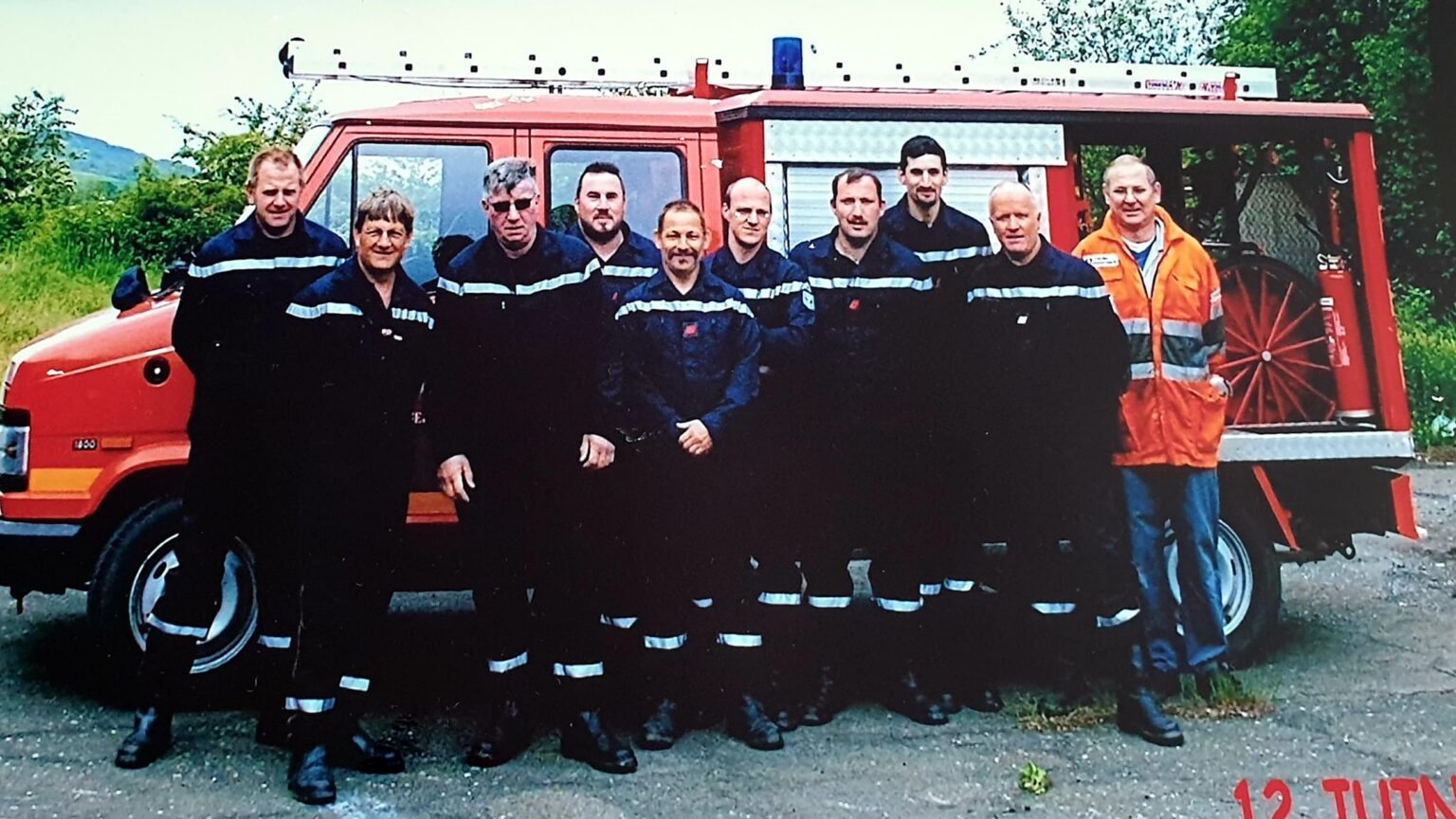 Dix hommes en tenue bleu marine de sapeur-pompier à bandes réfléchissantes, l'un d'eux en veste orange de haute visibilité, posent devant un fourgon rouge d'incendie.