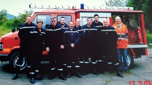 Dix hommes en tenue bleu marine de sapeur-pompier à bandes réfléchissantes, l'un d'eux en veste orange de haute visibilité, posent devant un fourgon rouge d'incendie.