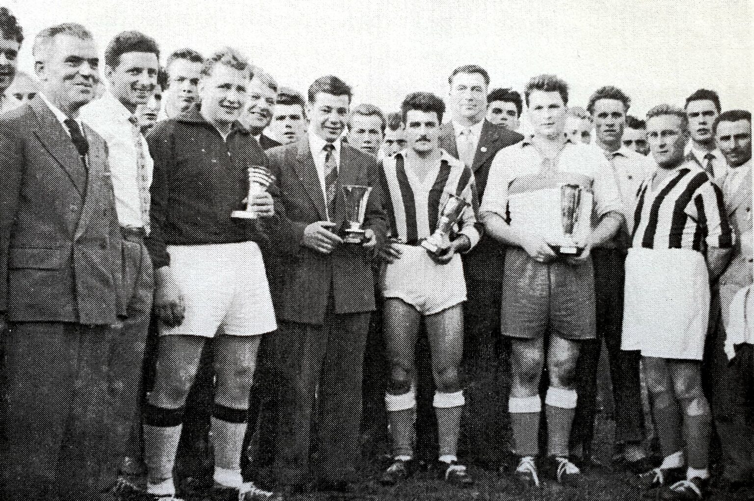 Photographie en noir et blanc de la remise des prix : les capitaines, en maillot sombre, rayé ou clair, tiennent chacun une petite coupe, entourés de dirigeants en costume cravate et des joueurs massés derrière eux.
