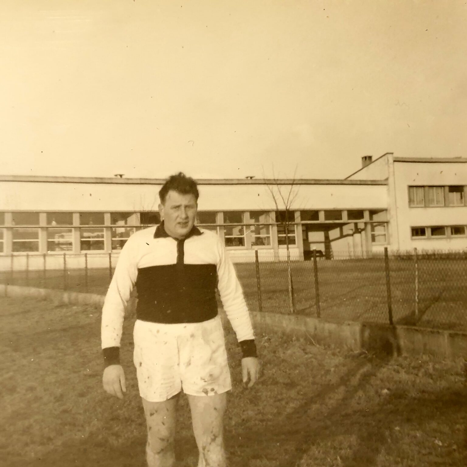 Un joueur seul au bord du terrain, maillot blanc barré d'une large bande noire et jambes maculées de boue ; derrière lui, la longue façade vitrée de l'école primaire.