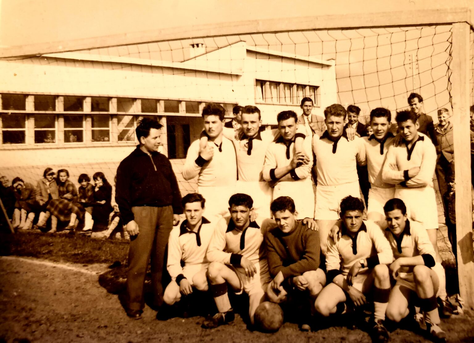 Photographie sépia : l'équipe posant dans les buts aux poteaux carrés, sept joueurs debout en maillot clair à col sombre et un dirigeant en blouson, cinq accroupis devant ; derrière, l'école et des spectateurs assis le long du grillage.