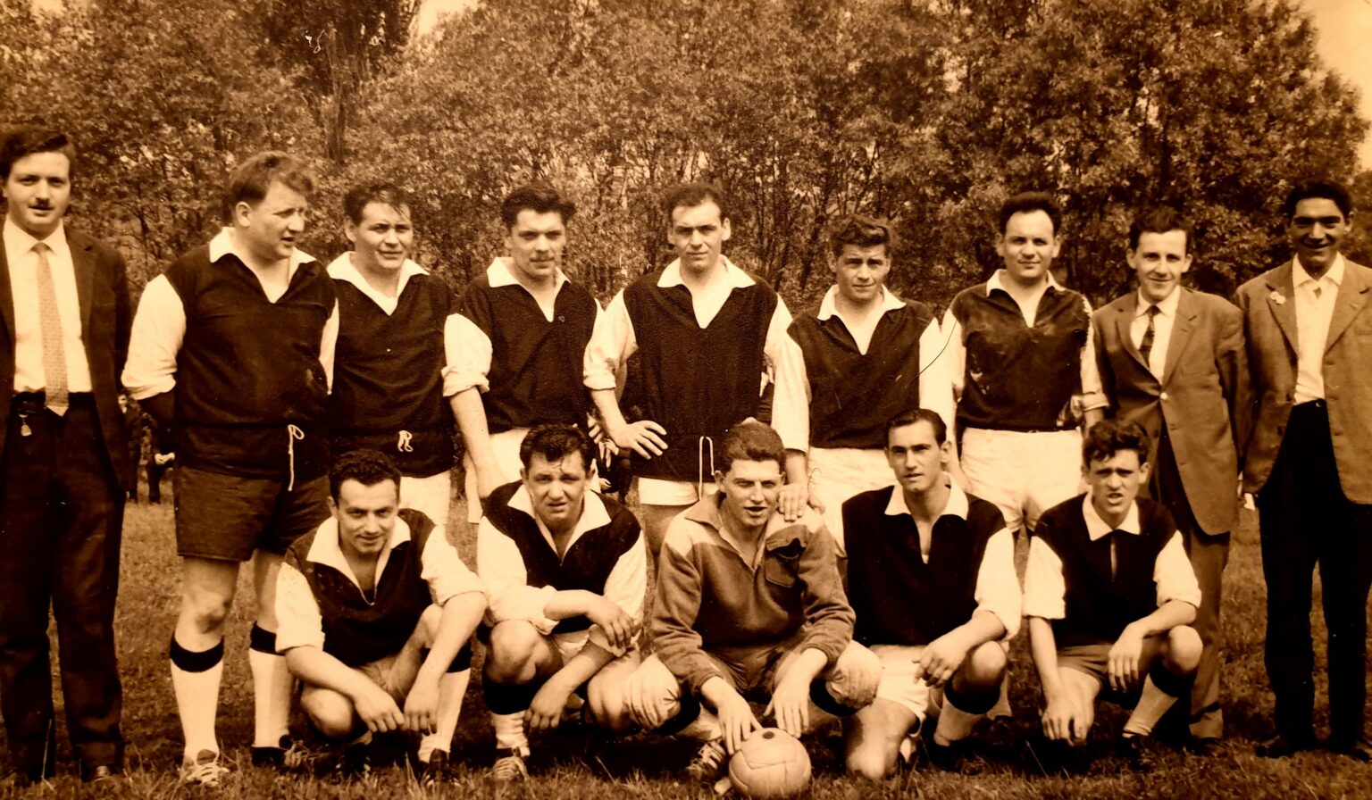 Photographie sépia : l'équipe en chemise blanche à manches retroussées sous un maillot sombre échancré, sept joueurs debout et deux dirigeants en costume aux extrémités, cinq joueurs accroupis devant, le ballon de cuir lacé posé au sol.