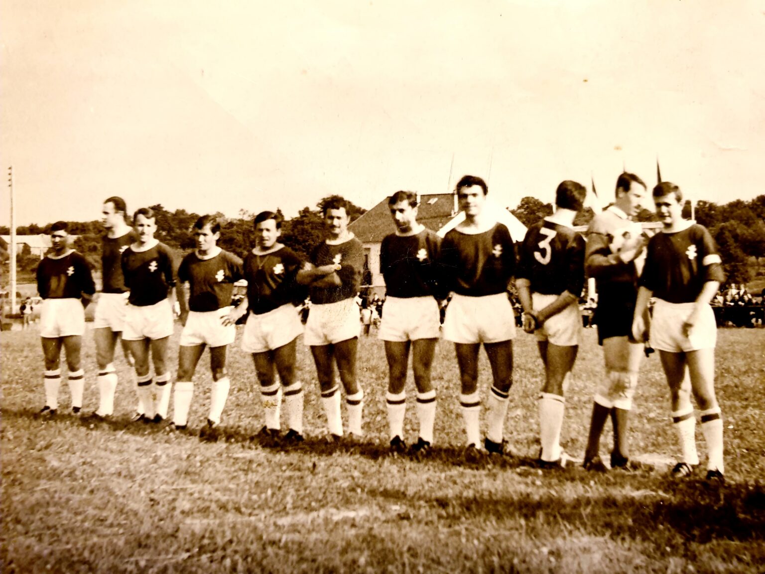 Photographie sépia : une équipe alignée sur le terrain avant le coup d'envoi, onze joueurs en maillot sombre à écusson et short blanc, devant le public massé le long de la ligne et une maison au fond.