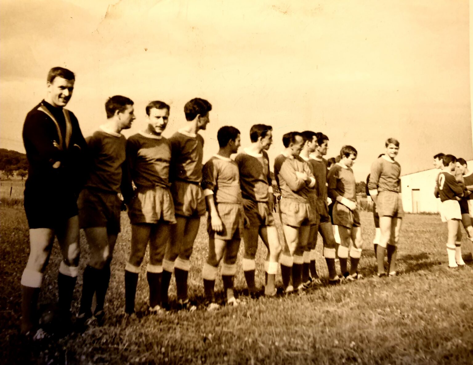 Photographie sépia : les joueurs alignés en file avant le match, le gardien tout en sombre au premier plan ; au loin, l'équipe adverse alignée face à eux.