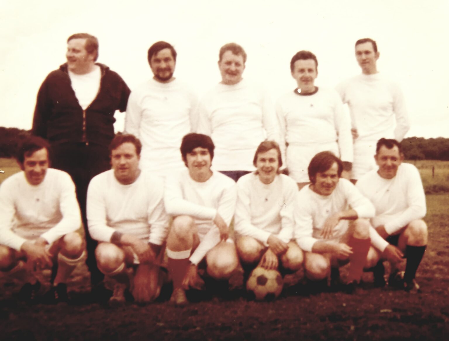 Photographie ancienne aux teintes sépia : l'équipe en maillot blanc à manches longues, quatre joueurs debout derrière un dirigeant en gilet foncé, et six accroupis devant, le ballon de cuir posé au sol.