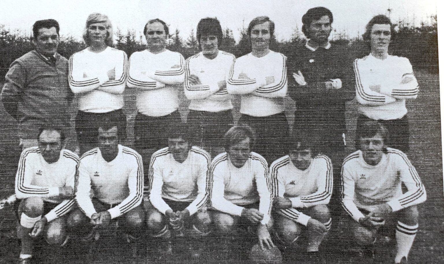 Photographie en noir et blanc de l'équipe en maillot blanc à trois bandes : six joueurs debout, bras croisés, encadrés d'un dirigeant en veste et d'un arbitre en noir, et six autres accroupis devant.