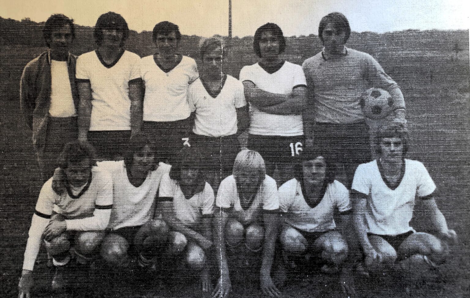 Photographie ancienne d'une équipe en maillot blanc : six hommes debout, dont un dirigeant en veste claire, et six joueurs accroupis devant, sur un terrain bordé de collines.