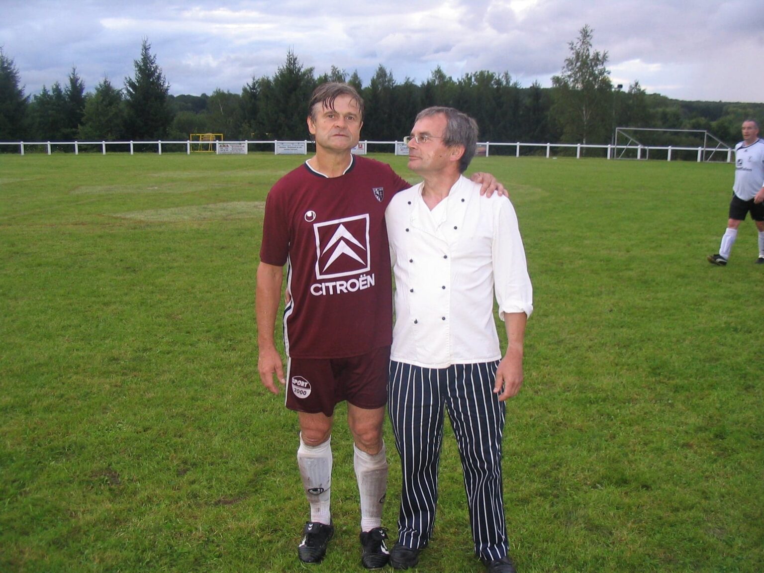 Deux hommes posent au milieu du terrain, le bras sur l'épaule : l'un en maillot grenat du FC Metz floqué « Citroën », l'autre en veste blanche et pantalon rayé de cuisinier.