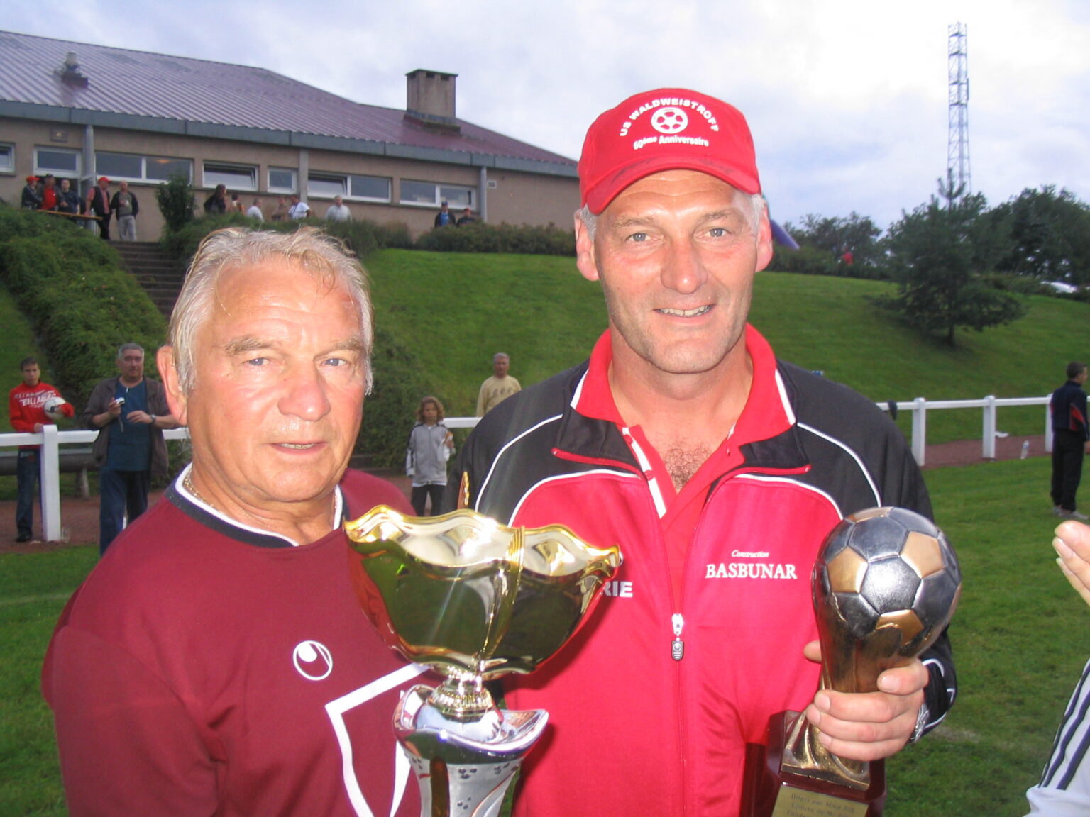 Deux hommes posent avec leurs trophées devant le talus du stade : l'un tient une grande coupe dorée, l'autre un ballon doré sur socle ; sa casquette rouge porte « US Waldweistroff, 60ème anniversaire ».