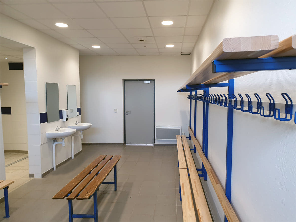 Un vestiaire neuf : des bancs de bois sur piètement bleu, une longue patère bleue à une trentaine de crochets, deux lavabos surmontés de miroirs et une porte au fond.