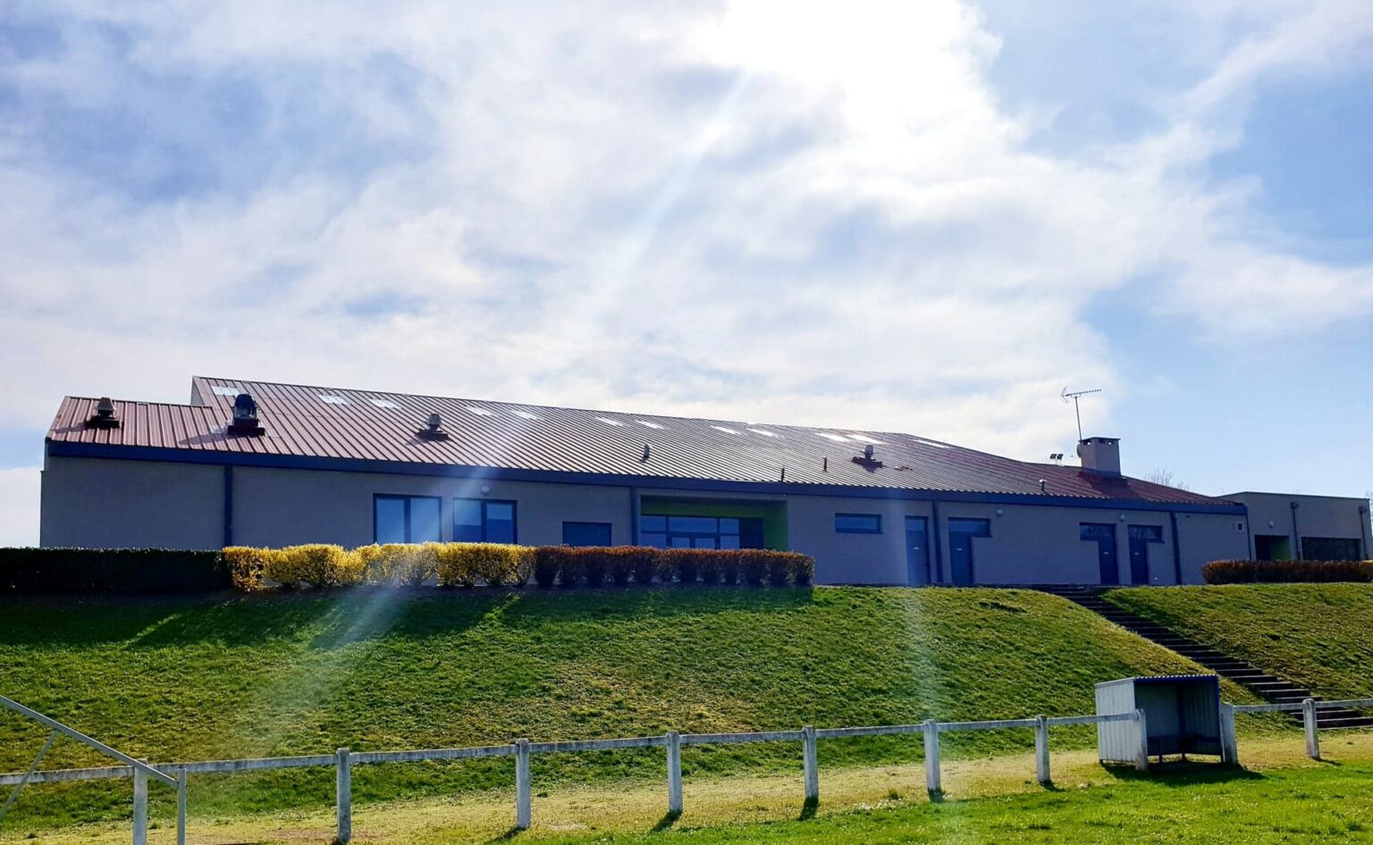 Le bâtiment du club, long et bas, en crépi gris sous une toiture métallique, domine le terrain depuis un talus engazonné ; en contrebas, la main courante blanche et un abri de touche.