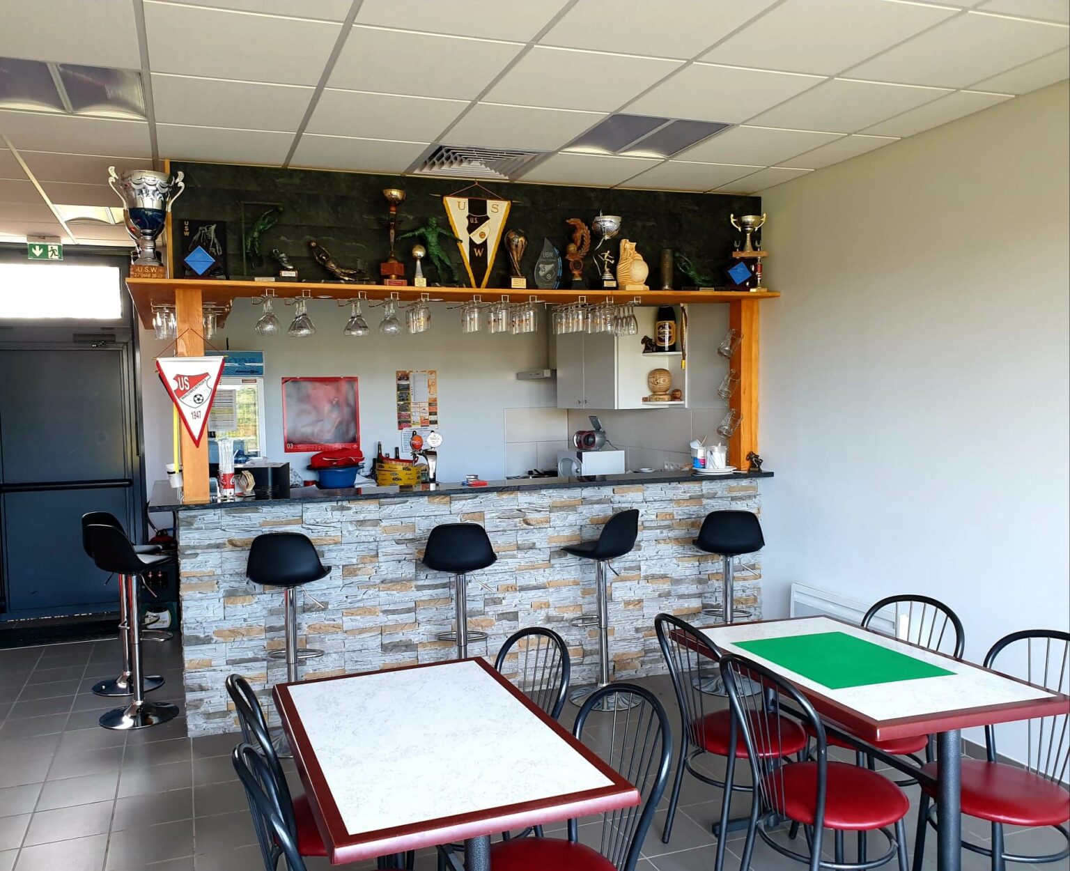 La salle du Club House : un comptoir habillé de parement de pierre, des tabourets noirs et des verres suspendus, surmontés d'une étagère garnie de coupes, de trophées et de fanions de l'U.S.W. ; devant, des tables et des chaises.