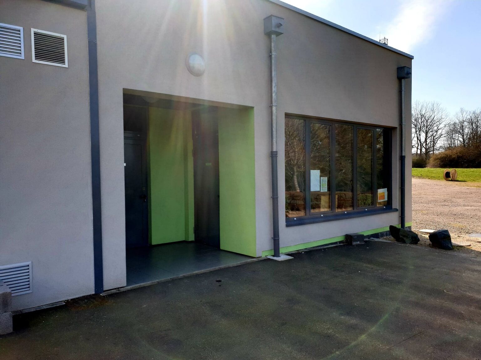 L'extérieur du Club House refait à neuf : un bâtiment en crépi gris, avec un porche ouvert peint en vert anis et une large baie vitrée.