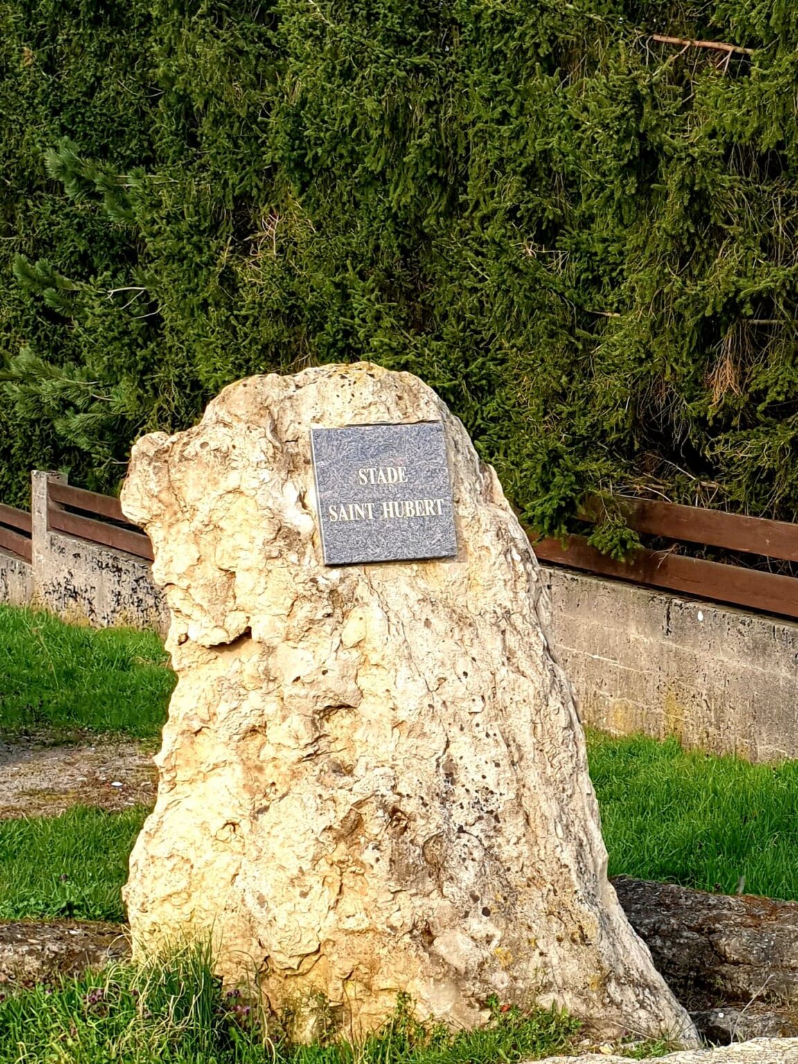 Un bloc de pierre dressé à l'entrée porte une plaque de granit gravée « STADE SAINT HUBERT », devant une haie de résineux.