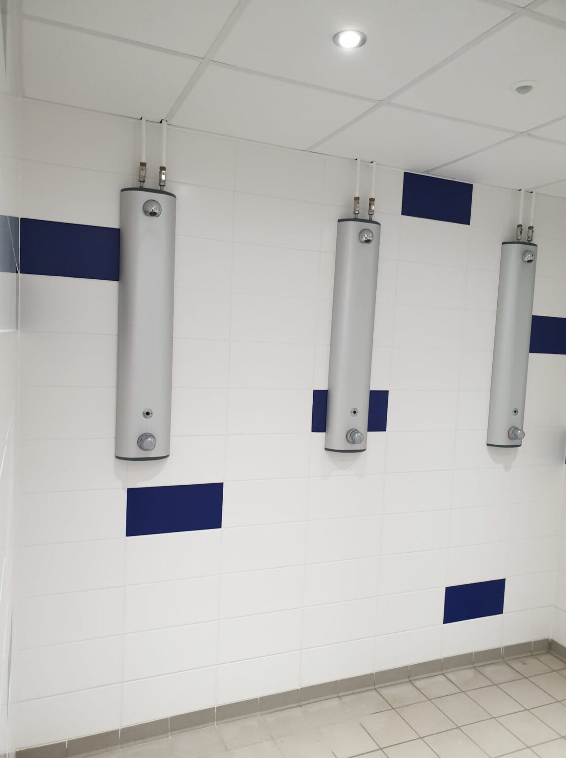 Les douches de l'autre vestiaire : trois colonnes de douche sur un carrelage blanc semé de carreaux bleu marine.